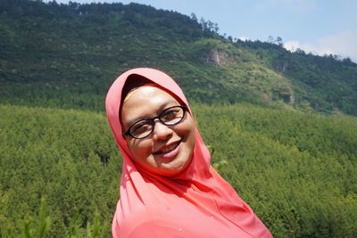 Portrait of cheerful young woman wearing hijab and eyeglasses against mountain during sunny day