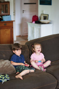 Siblings playing video game while sitting on sofa at home