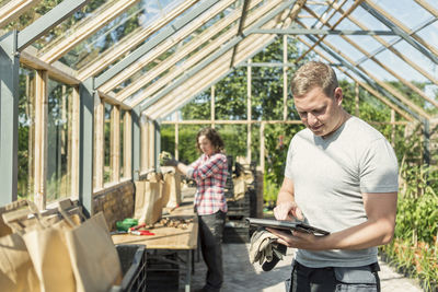 Man using digital tablet while woman working in greenhouse