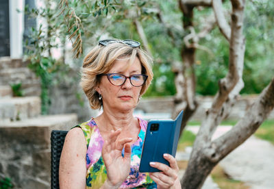 Senior woman sitting under olive tree in back yard, using mobile phone