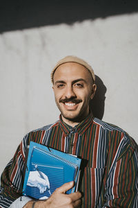 Portrait of smiling male adult student holding book standing in front of gray wall at college on sunny day