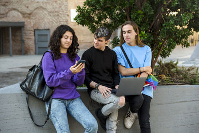 Young diverse people with lgbt rainbow flag using laptop and mobile outdoors.