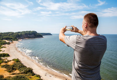 Rear view of man photographing sea through mobile phone