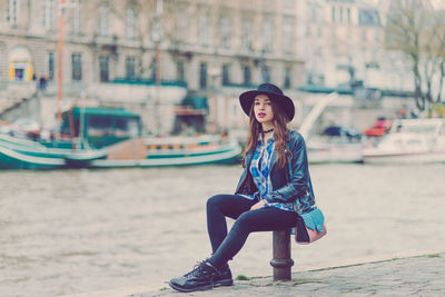 Full length portrait of beautiful woman sitting on bollard by seine river