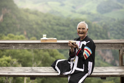 Portrait of senior woman holding coffee cup sitting on bench against landscape