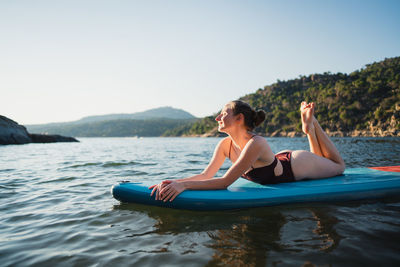 Full body of female in swimwear lying on paddleboard on rippling lake against shore with forest on sunny summer day