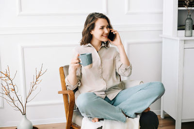 Cheerful young woman talking on smartphone, relaxing in armchair with drink. happy young woman