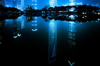 Illuminated buildings by river against blue sky at night