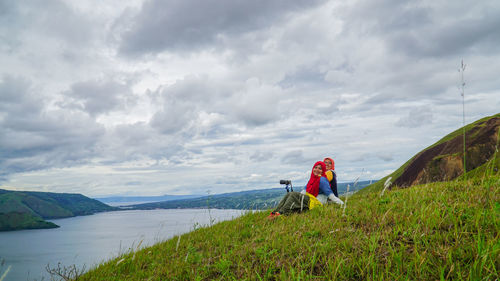 Female friends sitting on grassy hill by lake toba against sky