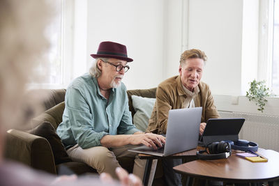 Senior men talking to each other while sitting in living room and using digital tablet and laptop to edit podcast