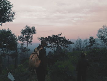 Rear view of man standing by trees against sky during sunset