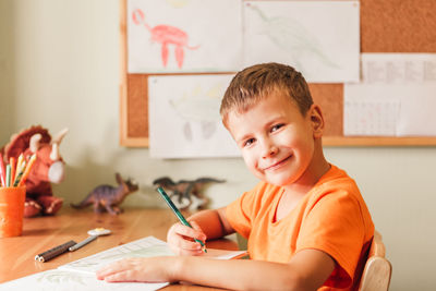 Cute child boy drawing dinosaurs on picture with colored pencils sitting by desk at his room