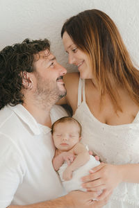 Cheerful hispanic man and woman with cute sleeping baby touching noses and smiling while hugging each other