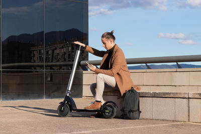 Young modern man sitting on a bench using mobile phone with his electric scooter with green  energy