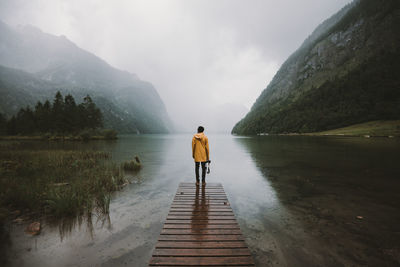 Rear view of man standing on pier at lake against mountains