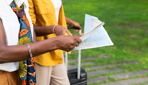 Crop young african american female friends in stylish clothes reading map while standing on street with suitcase during vacation