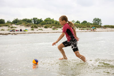 Full length of playful girl playing soccer ball in sea against sky