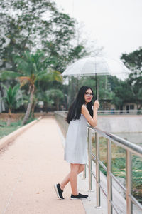 Portrait of young woman holding umbrella standing on footpath by railing in park