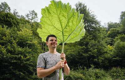 Portrait of young man holding  large burdock leaf while standing in forest