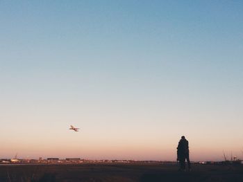 People standing in airfield as an airplane flies in sky at sunset