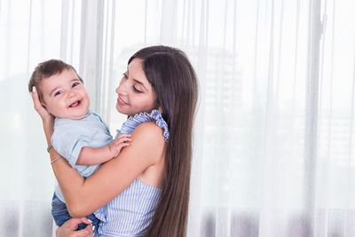 Side view of mother holding cheerful toddler son against curtain at home