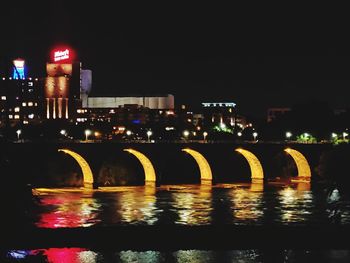 Illuminated bridge over river by buildings against sky at night