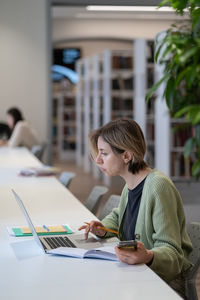 Focused female university teacher using laptop while sitting in quiet cozy library