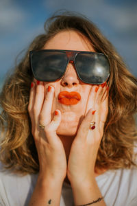 Stylish brown curly haired woman with red lipstick in trendy sunglasses looking at camera while squeezing face in palms