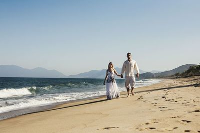 Couple holding hands while walking on shore at beach against sky