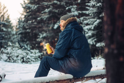 Side view of man having drink while sitting against trees during winter