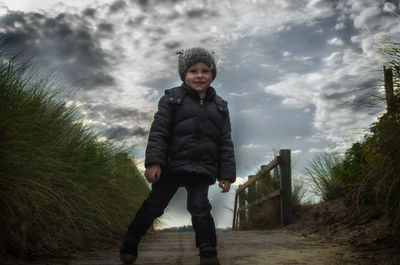 Portrait of boy standing on footbridge against cloudy sky
