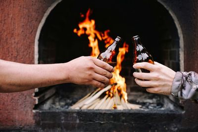 Close-up of hands holding bottles of beer in front of wood fire