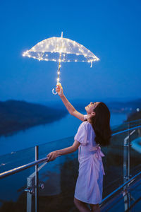 Young woman holding illuminated umbrella standing by railing during dusk