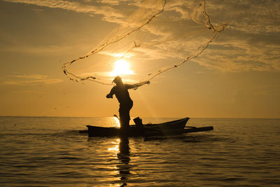 Silhouette fisherman throwing fishing net in sea against sky during sunset