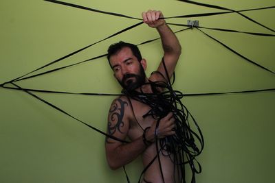 Naked man looking away while holding cables against wall