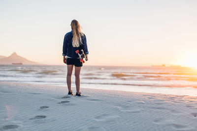 Rear view of young woman with skateboard standing at beach against clear sky during sunset