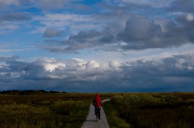 Rear view of person riding bicycle on footpath against cloudy sky