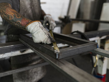 Unrecognizable male welder in dirty gloves and apron measuring frame with yellow metal tape and working at workbench in garage