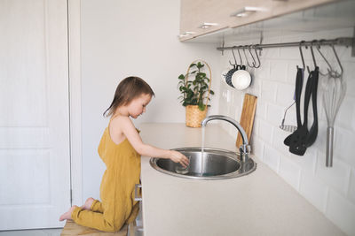 European child of four years old in yellow jumpsuit washes his hands in real bright kitchen