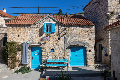 A weathered stone house with vibrant blue doors and shutters sits on a sunlit mediterranean street.