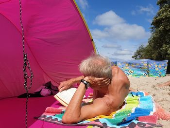 Senior shirtless man reading book while lying down on beach by tent