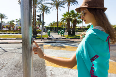 Side view of young woman pressing button in pole on city street