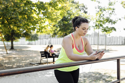 Young woman in sportswear using smart phone while leaning on railing at park