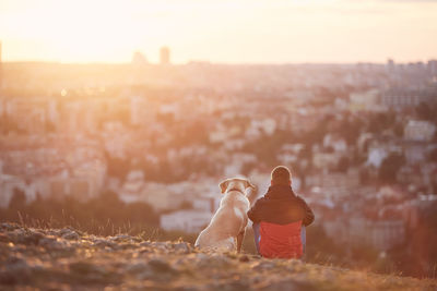 Rear view of young man with dog at sunrise. pet owner sitting with his dog on hill against city. 