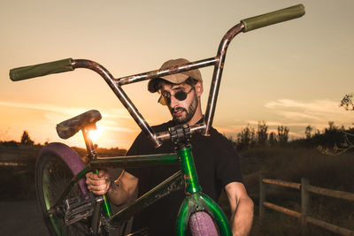 Portrait of man holding bicycle on road against sky during sunset