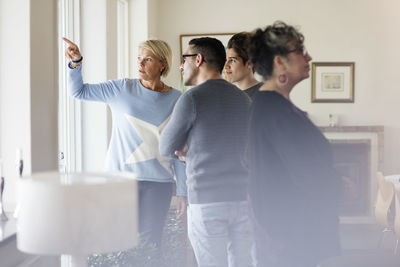 Mature woman pointing and showing to friends through window while standing in living room