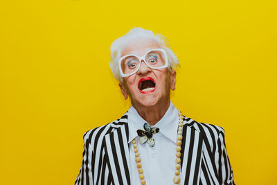 Portrait of angry senior woman wearing eyeglasses against yellow background