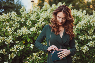 Smiling young woman holding purse while standing against plants