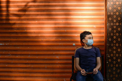 Boy wearing mask looking away while sitting against shutter