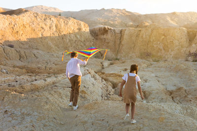 A girl and a boy are running around a sandy quarry with a bright kite.